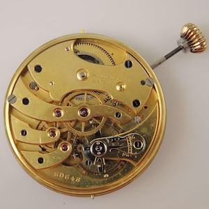 May include: A gold-colored pocket watch movement with intricate gears and a winding stem. The movement is labeled "Swiss Made" and has the number "60648" printed on it.