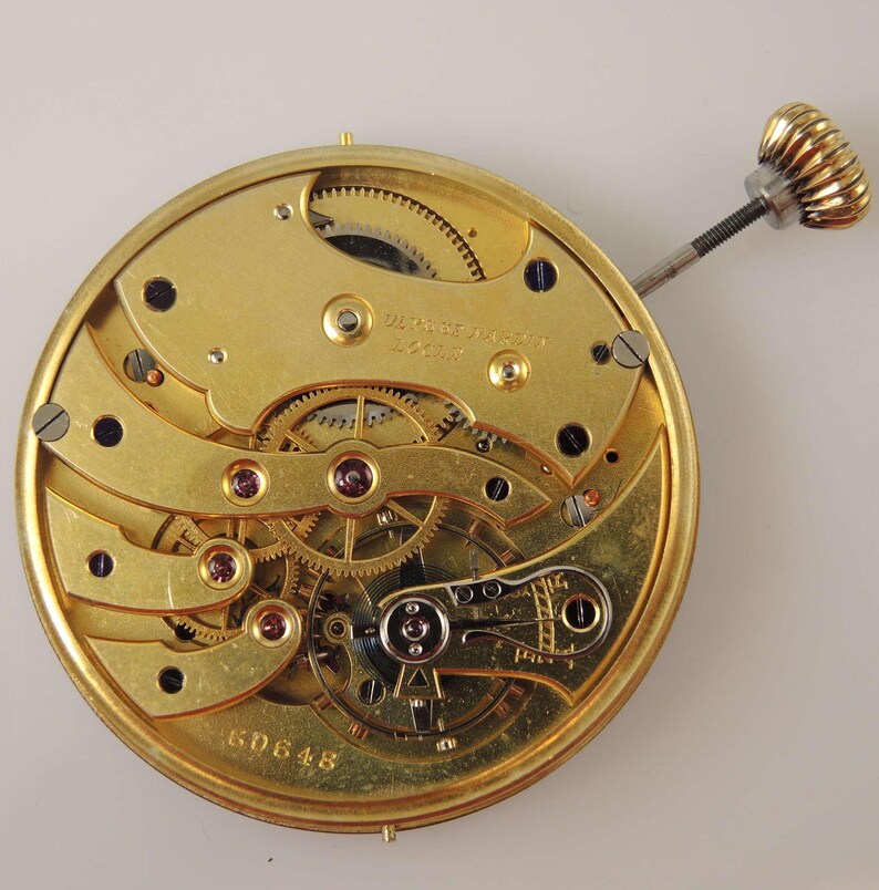 May include: A gold-colored pocket watch movement with intricate gears and a winding stem. The movement is labeled "Swiss Made" and has the number "60648" printed on it.