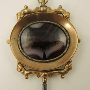 May include: Antique gold-toned pendant with an oval, banded agate stone. The stone is framed by a detailed, ornate setting with decorative elements. A small loop and a winding key are attached.