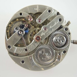 Op de afbeelding: Close-up van een zilveren zakhorloge-uurwerk met ingewikkelde tandwielen en een handtekening die "Alfred Mannhardt, Genf" luidt.