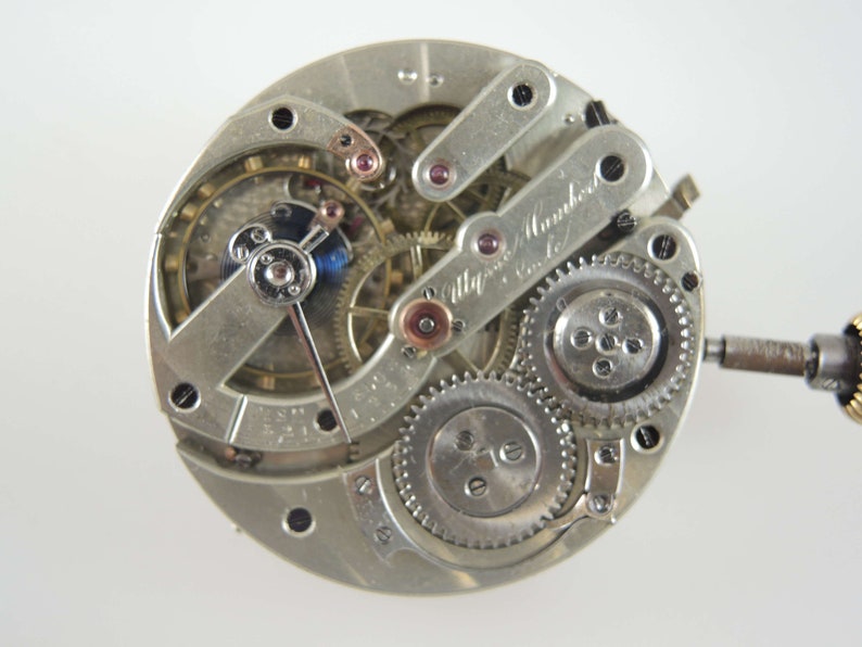 Op de afbeelding: Close-up van een zilveren zakhorloge-uurwerk met ingewikkelde tandwielen en een handtekening die "Alfred Mannhardt, Genf" luidt.