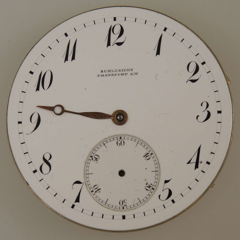 May include: A white pocket watch face with black Roman numeral hour markers and black Arabic numeral minute markers. The watch face has a smaller white subdial with black Arabic numeral markers. The watch face also has the text "SCHLESICKY FRANKFURT A/M".