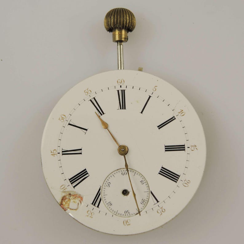 May include: Antique pocket watch face with a white dial, black Roman numerals, and gold hands. The watch has a small seconds dial and a decorative brass crown. The face shows some signs of age, adding to its vintage charm.