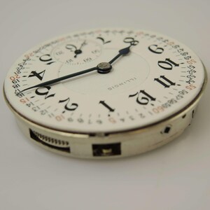 May include: A close-up of a vintage watch face with black numerals on a white background. The watch face has a smaller subdial and black hands. The word "ILLINOIS" is printed on the face. A second hand dial is visible.