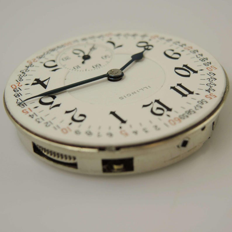May include: A close-up of a vintage watch face with black numerals on a white background. The watch face has a smaller subdial and black hands. The word "ILLINOIS" is printed on the face. A second hand dial is visible.