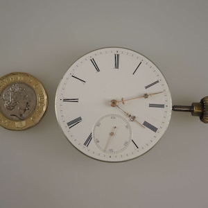 May include: A white pocket watch with Roman numeral hour markers and a smaller subdial. The watch has a gold-colored winding stem and is about the size of a British pound coin.