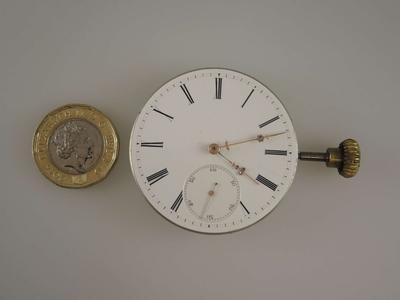 May include: A white pocket watch with Roman numeral hour markers and a smaller subdial. The watch has a gold-colored winding stem and is about the size of a British pound coin.