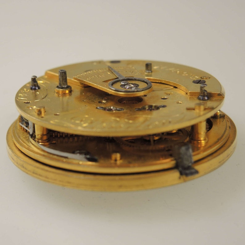 Puede incluir: Primer plano de un movimiento de reloj de bolsillo de tono dorado con engranajes intrincados y un mecanismo de cuerda. El reloj est&aacute; desmontado y se pueden ver las partes internas.