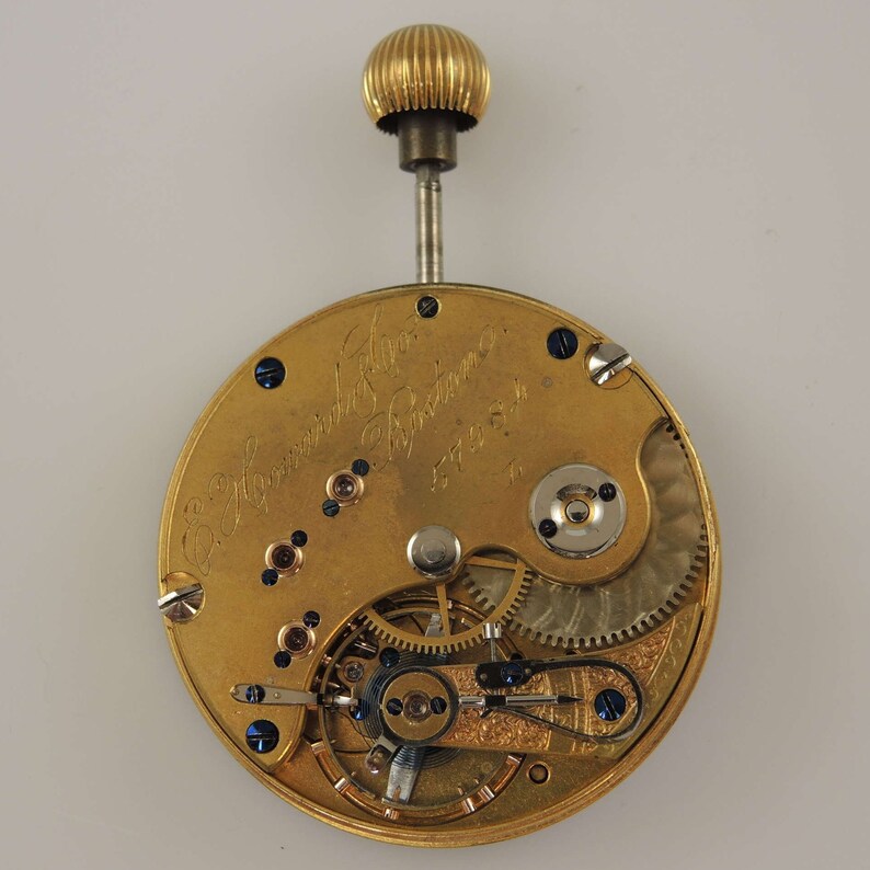 Puede incluir: Un movimiento de reloj de bolsillo de oro con la inscripci&oacute;n "E. Howard & Co. Boston" y el n&uacute;mero "57454". El movimiento est&aacute; abierto y muestra los intrincados engranajes y resortes.