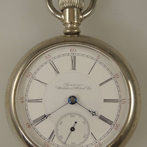 Pode incluir: Um relógio de bolso prateado com numerais romanos no mostrador. O mostrador do relógio diz "American Waltham Watch Co."