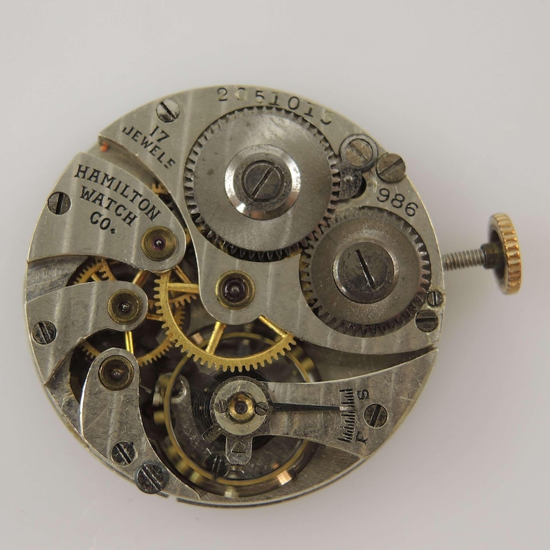 K&ouml;nnte beinhalten: Nahaufnahme eines Vintage-Uhrwerks der Hamilton Watch Co. Der silberfarbene Mechanismus zeigt komplizierte Zahnr&auml;der, Schrauben und den Text "17 Jewels". Eine goldfarbene Krone ist rechts zu sehen. Die Nummer "986" ist auf das Uhrwerk gestempelt.