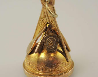 May include: A gold-coloured antique pendant with intricate detailing. The pendant has a triangular top with engraved patterns and a circular base with floral engravings. A small emblem with a rose and triangle is visible.