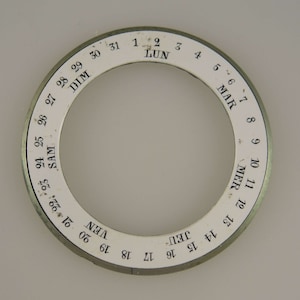 Könnte beinhalten: Kreisförmiges weiß-silbernes Zifferblatt mit schwarzen Zahlen und Text. Das Zifferblatt zeigt die Tage des Monats von 1 bis 31 und die Wochentage auf Französisch an. Eine Vintage-Uhrenkomponente.