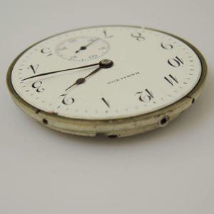 May include: Close-up of a vintage Hamilton pocket watch face. The white face features black numerals, a smaller subdial, and black hands. The word "HAMILTON" is printed in black. The watch face has a silver-colored metal rim.