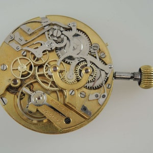 May include: A close-up of a gold-toned pocket watch movement with intricate gears and levers. The watch is disassembled and the inner workings are visible. The watch has a winding key and a small knob on the side.