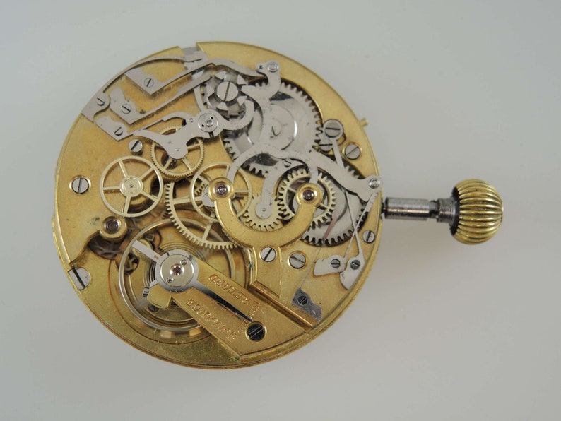 May include: A close-up of a gold-toned pocket watch movement with intricate gears and levers. The watch is disassembled and the inner workings are visible. The watch has a winding key and a small knob on the side.