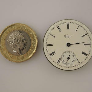 May include: An antique Elgin pocket watch face with black Roman numerals on an off-white background. The watch face includes a smaller seconds dial and black hands. A gold coin is next to the watch face.