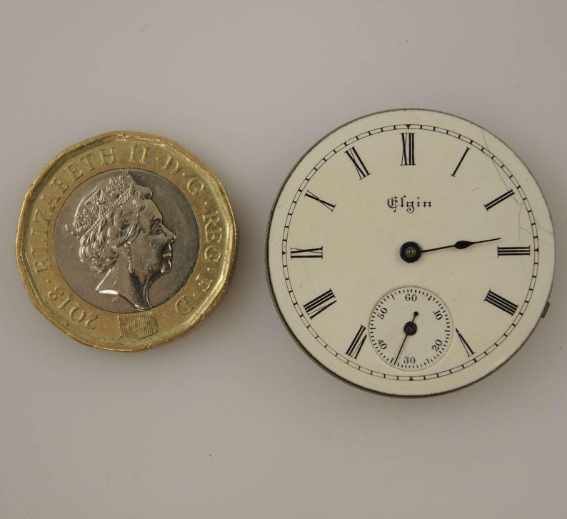 May include: An antique Elgin pocket watch face with black Roman numerals on an off-white background. The watch face includes a smaller seconds dial and black hands. A gold coin is next to the watch face.