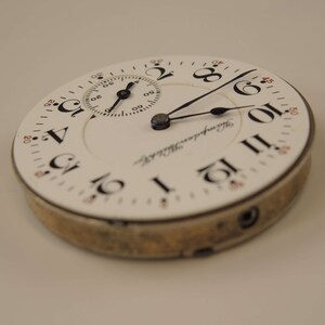 K&ouml;nnte beinhalten: Eine wei&szlig;e Taschenuhr mit schwarzen r&ouml;mischen Ziffern f&uuml;r die Stunden und schwarzen Zeigern. Das Zifferblatt der Uhr hat ein kleineres, kreisf&ouml;rmiges Hilfszifferblatt mit schwarzen Zeigern und schwarzen arabischen Ziffern. Auf dem Zifferblatt der Uhr steht auch der Text "Hampden Watch Co.".