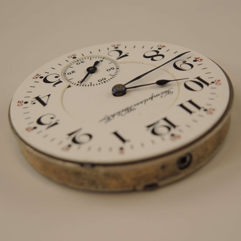K&ouml;nnte beinhalten: Eine wei&szlig;e Taschenuhr mit schwarzen r&ouml;mischen Ziffern f&uuml;r die Stunden und schwarzen Zeigern. Das Zifferblatt der Uhr hat ein kleineres, kreisf&ouml;rmiges Hilfszifferblatt mit schwarzen Zeigern und schwarzen arabischen Ziffern. Auf dem Zifferblatt der Uhr steht auch der Text "Hampden Watch Co.".