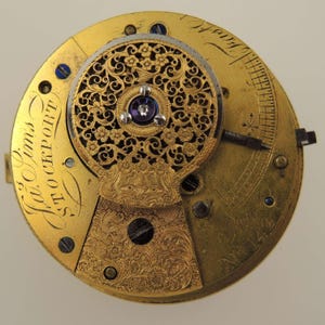 May include: Close-up of a vintage watch movement with intricate gold filigree and floral designs. The watch mechanism features a central jewel and engraved details. The text "W. Sims STOCKPORT" is visible on the gold-toned surface.