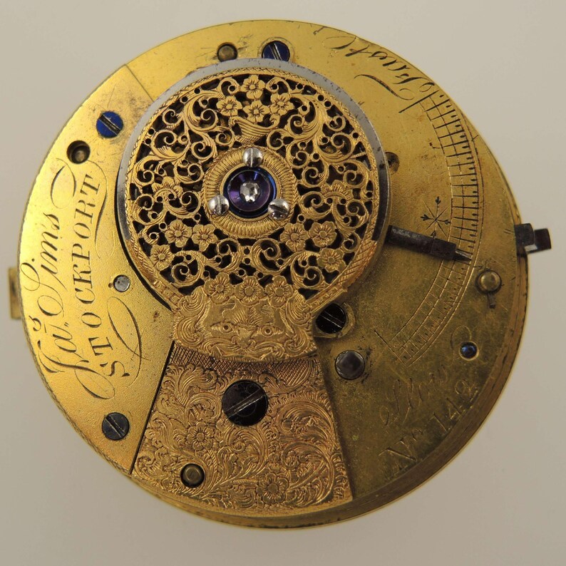 May include: Close-up of a vintage watch movement with intricate gold filigree and floral designs. The watch mechanism features a central jewel and engraved details. The text "W. Sims STOCKPORT" is visible on the gold-toned surface.