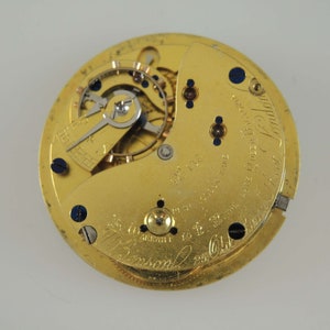 Op de afbeelding: Close-up van een goudkleurig uurwerk met ingewikkelde tandwielen en componenten. Het horlogemechanisme heeft kleine schroeven en blauwe accenten. De wijzerplaat heeft gegraveerde tekst, waaronder "J. Benson" en "London".