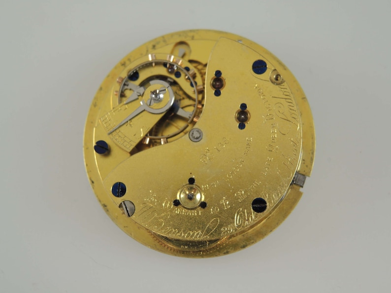 Op de afbeelding: Close-up van een goudkleurig uurwerk met ingewikkelde tandwielen en componenten. Het horlogemechanisme heeft kleine schroeven en blauwe accenten. De wijzerplaat heeft gegraveerde tekst, waaronder "J. Benson" en "London".