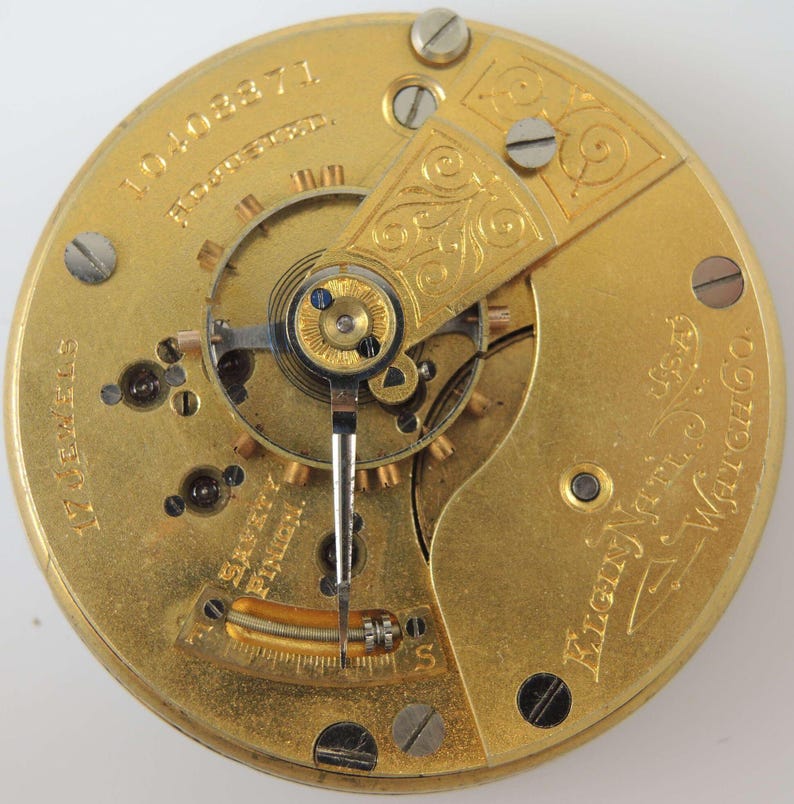 K&ouml;nnte beinhalten: Nahaufnahme des Innenlebens einer goldfarbenen Taschenuhr. Das Zifferblatt der Uhr ist mit dem Text "Elgin National Watch Co.", "14 Jewels" und "10403371" graviert.