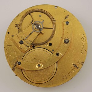 May include: Close-up of a gold-colored pocket watch movement with intricate gears and a lever. The watch is labeled "A.L. Adams" and "Geneva" on the back.