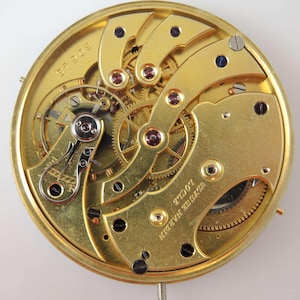 May include: A gold-colored pocket watch movement with intricate gears and a winding mechanism. The watch is labeled "Ulysse Nardin" and "Locle" on the back.