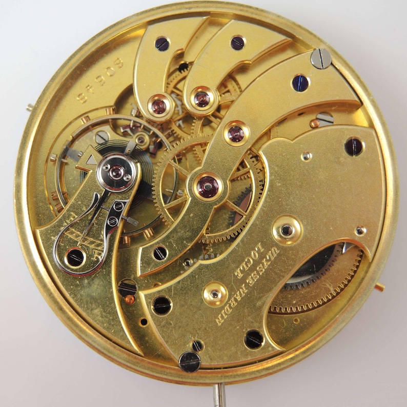 May include: A gold-colored pocket watch movement with intricate gears and a winding mechanism. The watch is labeled "Ulysse Nardin" and "Locle" on the back.
