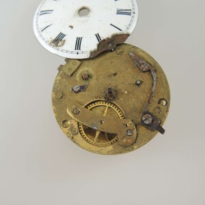K&ouml;nnte beinhalten: Eine zerlegte antike Taschenuhr mit wei&szlig;em Zifferblatt und vergoldeten Zahnr&auml;dern. Dem Zifferblatt fehlt ein Teil des &auml;u&szlig;eren Randes.