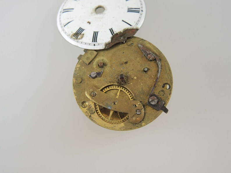 K&ouml;nnte beinhalten: Eine zerlegte antike Taschenuhr mit wei&szlig;em Zifferblatt und vergoldeten Zahnr&auml;dern. Dem Zifferblatt fehlt ein Teil des &auml;u&szlig;eren Randes.