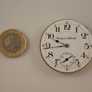 K&ouml;nnte beinhalten: Eine wei&szlig;e Taschenuhr mit schwarzen Zeigern und r&ouml;mischen Ziffern. Das Zifferblatt der Uhr tr&auml;gt den Schriftzug "Hampden Watch Co.".