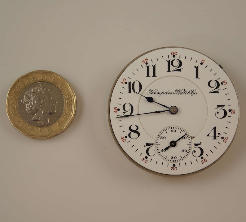 K&ouml;nnte beinhalten: Eine wei&szlig;e Taschenuhr mit schwarzen Zeigern und r&ouml;mischen Ziffern. Das Zifferblatt der Uhr tr&auml;gt den Schriftzug "Hampden Watch Co.".