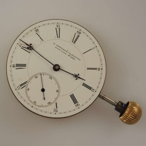 Puede incluir: Un reloj de bolsillo blanco con marcadores de hora en n&uacute;meros romanos negros y marcadores de minutos negros. La esfera del reloj tiene el texto "E Howard & Co Boston Pat'd Feb 25, 1868".