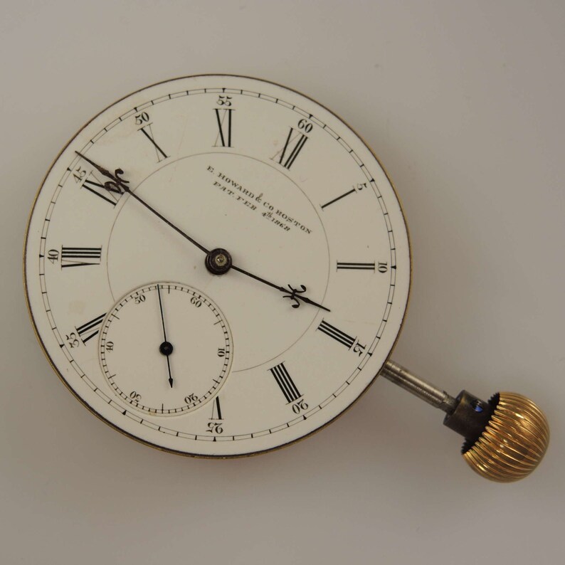 Puede incluir: Un reloj de bolsillo blanco con marcadores de hora en n&uacute;meros romanos negros y marcadores de minutos negros. La esfera del reloj tiene el texto "E Howard & Co Boston Pat'd Feb 25, 1868".