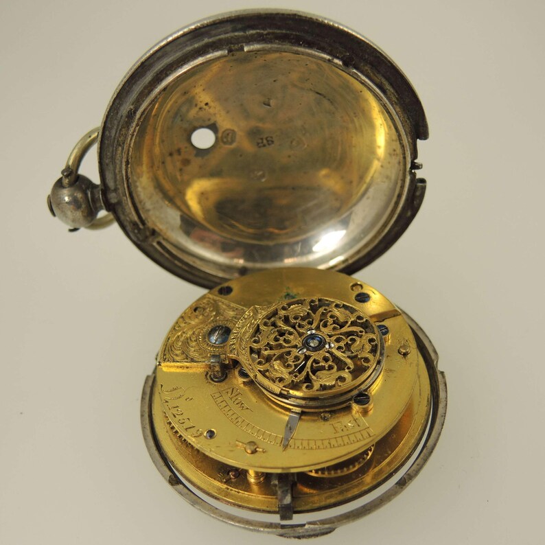 May include: An antique pocket watch with the case open, revealing the intricate gold-colored inner workings. The silver-colored case is partially open, showcasing the watch's detailed mechanism and engraved components. The watch is a vintage item.