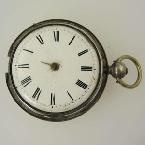 May include: A vintage pocket watch with a white face and black Roman numerals. The watch has a silver-colored casing and a gold-colored ring for attaching a chain. The watch face shows some signs of age.
