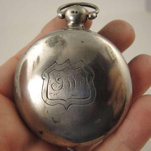 Könnte beinhalten: Eine silberne Taschenuhr mit einer schildförmigen Gravur. Die Uhr hat einen kreisförmigen Körper mit einer Schlaufe und zwei kleinen Knöpfen oben. Das Schilddesign zeigt eine verzierte Initiale oder ein Monogramm. Die Uhr wird in einer Hand gehalten.