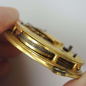 May include: Close-up of a gold watch mechanism. The internal components are visible, including gears and a textured wheel. The watch appears to be vintage, showcasing detailed craftsmanship. The mechanism is likely from a pocket watch.