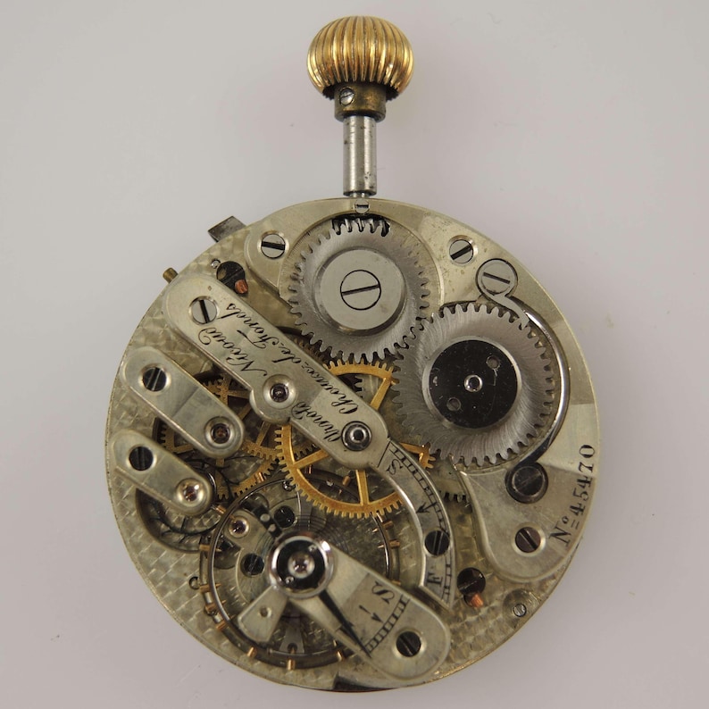 May include: Close-up of a vintage watch movement with intricate gears and mechanisms. The watch has a gold-colored crown and a silver-toned body. The text "Chaux de Fonds" is visible on a metal component, along with the number "45470".