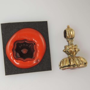 May include: A gold-colored antique wax seal with a decorative handle next to a red wax impression on a black square. The wax impression has the word "STAND" in the center.