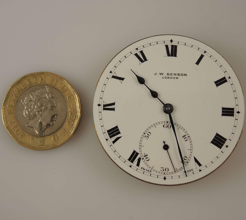 Puede incluir: Una esfera de reloj de bolsillo blanca con marcadores de hora en n&uacute;meros romanos negros y agujas negras. La esfera del reloj tiene un anillo exterior negro con marcas para segundos y minutos. La esfera del reloj tambi&eacute;n tiene el texto "J.W. BENSON LONDON" impreso en ella.