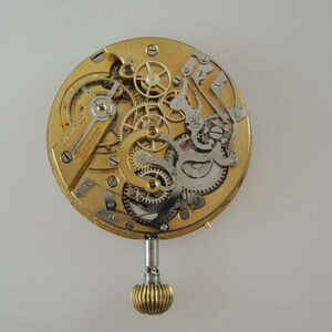 May include: A gold-colored watch movement with intricate gears and a winding stem. The movement is exposed, showing the inner workings of the watch.