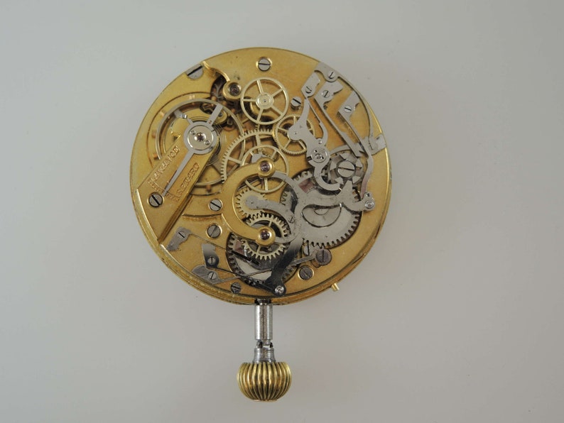 May include: A gold-colored watch movement with intricate gears and a winding stem. The movement is exposed, showing the inner workings of the watch.