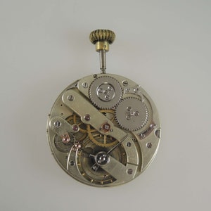 May include: A silver pocket watch movement with exposed gears and a winding stem. The watch is disassembled and shows the inner workings of the timepiece.