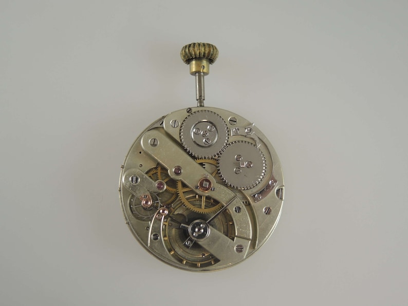 May include: A silver pocket watch movement with exposed gears and a winding stem. The watch is disassembled and shows the inner workings of the timepiece.