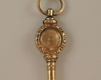May include: A gold-toned pocket watch key with a decorative round head and a loop at the top.